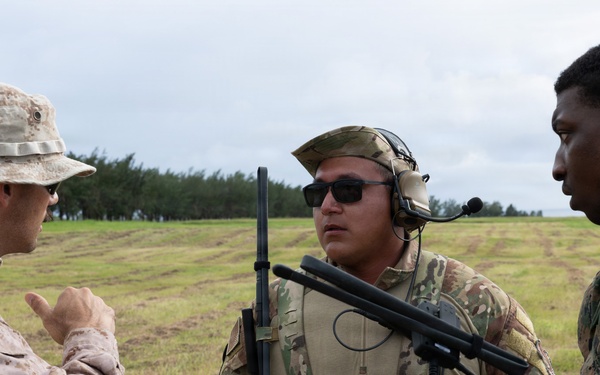 Three military personnel are engaged in a discussion in an open field environment. Two individuals wear U.S. Marine Corps uniforms (desert MARPAT and woodland MARPAT patterns), while the center individual wears Army MultiCam/OCP pattern with tactical communications gear. The setting appears to be a training exercise or operational briefing in a grassy field.