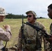 Three military personnel are engaged in a discussion in an open field environment. Two individuals wear U.S. Marine Corps uniforms (desert MARPAT and woodland MARPAT patterns), while the center individual wears Army MultiCam/OCP pattern with tactical communications gear. The setting appears to be a training exercise or operational briefing in a grassy field.