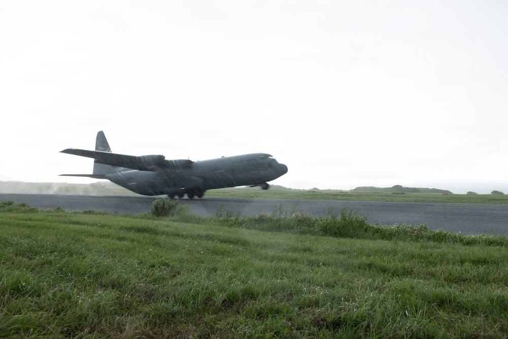 A military turboprop transport aircraft, consistent with a C-130 Hercules, is captured during takeoff or landing on a wet runway in rainy conditions. The aircraft is at low altitude with its landing gear still deployed, kicking up spray and mist from the wet tarmac. The surrounding terrain is flat and grassy under an overcast sky.
