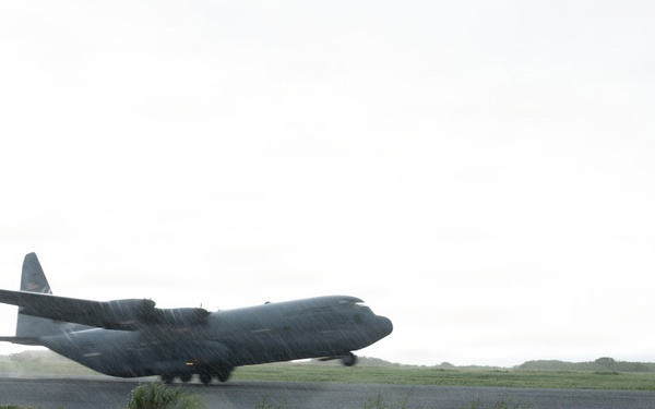 A military turboprop transport aircraft, consistent with a C-130 Hercules, is captured during takeoff or landing on a wet runway in rainy conditions. The aircraft is at low altitude with its landing gear still deployed, kicking up spray and mist from the wet tarmac. The surrounding terrain is flat and grassy under an overcast sky.