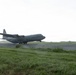 A military turboprop transport aircraft, consistent with a C-130 Hercules, is captured during takeoff or landing on a wet runway in rainy conditions. The aircraft is at low altitude with its landing gear still deployed, kicking up spray and mist from the wet tarmac. The surrounding terrain is flat and grassy under an overcast sky.