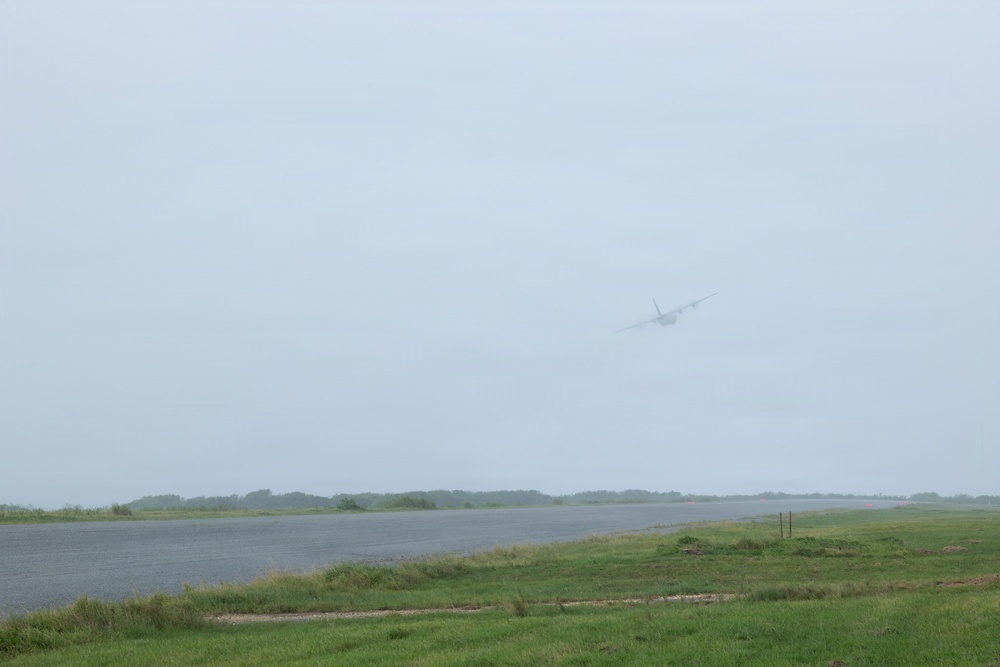 A large aircraft is captured in low visibility conditions, appearing to take off or fly at low altitude over what appears to be an airport runway. The scene is shrouded in overcast, hazy weather conditions that significantly reduce image clarity. Green grass borders the paved runway surface in the foreground.