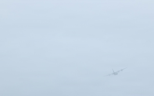 A large aircraft is captured in low visibility conditions, appearing to take off or fly at low altitude over what appears to be an airport runway. The scene is shrouded in overcast, hazy weather conditions that significantly reduce image clarity. Green grass borders the paved runway surface in the foreground.