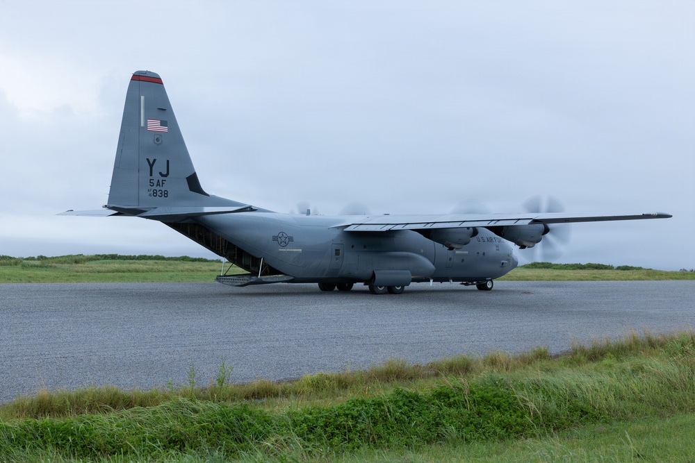 A U.S. Air Force C-130 Hercules military transport aircraft is parked or taxiing on a tarmac apron. The aircraft bears tail code 'YJ' indicating assignment to 5th Air Force, with serial number 838 visible. The rear cargo ramp appears to be open or lowered, and the propellers are spinning, suggesting the engines are running.