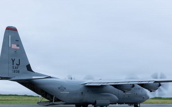A U.S. Air Force C-130 Hercules military transport aircraft is parked or taxiing on a tarmac apron. The aircraft bears tail code 'YJ' indicating assignment to 5th Air Force, with serial number 838 visible. The rear cargo ramp appears to be open or lowered, and the propellers are spinning, suggesting the engines are running.