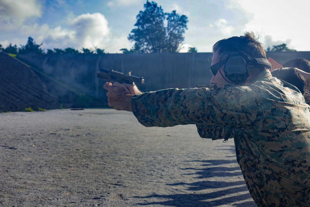 A person in military digital camouflage uniform is firing a semi-automatic pistol at an outdoor shooting range. The shooter is wearing hearing protection (earmuffs) and eye protection, using a two-handed grip stance. The image appears to capture an active firing moment, possibly during a military training exercise.