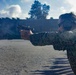 A person in military digital camouflage uniform is firing a semi-automatic pistol at an outdoor shooting range. The shooter is wearing hearing protection (earmuffs) and eye protection, using a two-handed grip stance. The image appears to capture an active firing moment, possibly during a military training exercise.