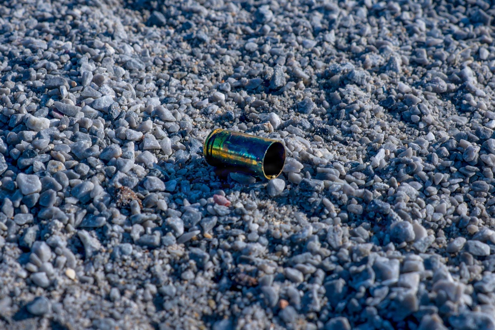 The image shows a single spent brass cartridge casing lying on a gravel surface. The casing displays an iridescent blue-green heat discoloration, indicating it has been fired. The surrounding ground consists of crushed gray gravel and small stones.