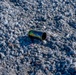 The image shows a single spent brass cartridge casing lying on a gravel surface. The casing displays an iridescent blue-green heat discoloration, indicating it has been fired. The surrounding ground consists of crushed gray gravel and small stones.