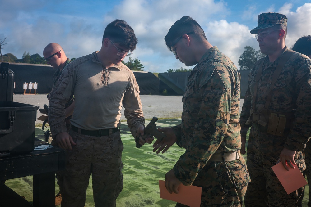 Military personnel, appearing to be U.S. Marines based on MARPAT camouflage uniforms, are gathered at an outdoor shooting range. Two central figures are examining a handgun, likely during a weapons inspection or qualification exercise. Additional personnel are visible in the background near shooting targets.