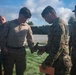 Military personnel, appearing to be U.S. Marines based on MARPAT camouflage uniforms, are gathered at an outdoor shooting range. Two central figures are examining a handgun, likely during a weapons inspection or qualification exercise. Additional personnel are visible in the background near shooting targets.