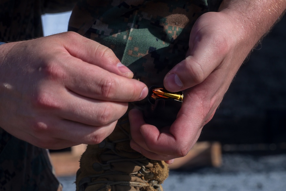 A person wearing woodland digital camouflage uniform is handling a single brass cartridge/bullet with both hands. The image is a close-up shot focusing on the hands and the ammunition. The setting appears to be an outdoor military training or range environment.