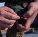 A person wearing woodland digital camouflage uniform is handling a single brass cartridge/bullet with both hands. The image is a close-up shot focusing on the hands and the ammunition. The setting appears to be an outdoor military training or range environment.