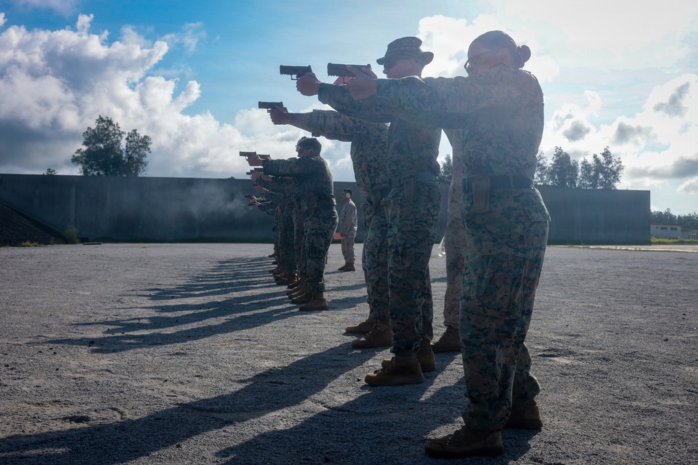 A line of military personnel in camouflage uniforms are conducting pistol qualification or training at an outdoor shooting range. The shooters are aligned in a row, all extending their sidearms in a two-handed shooting stance toward an unseen target downrange. A supervisor or range safety officer in lighter-colored clothing is visible in the background observing the exercise.
