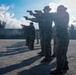 A line of military personnel in camouflage uniforms are conducting pistol qualification or training at an outdoor shooting range. The shooters are aligned in a row, all extending their sidearms in a two-handed shooting stance toward an unseen target downrange. A supervisor or range safety officer in lighter-colored clothing is visible in the background observing the exercise.