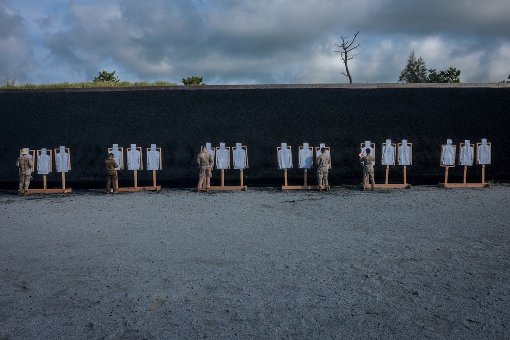 A military or law enforcement shooting range is depicted with personnel in camouflage uniforms standing at a target line inspecting or scoring paper silhouette targets. The range features a large earthen or rubber backstop berm running the full width of the image. Approximately five to six individuals are spaced along the target line, each positioned near clusters of humanoid paper targets mounted on wooden stands.