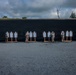 A military or law enforcement shooting range is depicted with personnel in camouflage uniforms standing at a target line inspecting or scoring paper silhouette targets. The range features a large earthen or rubber backstop berm running the full width of the image. Approximately five to six individuals are spaced along the target line, each positioned near clusters of humanoid paper targets mounted on wooden stands.