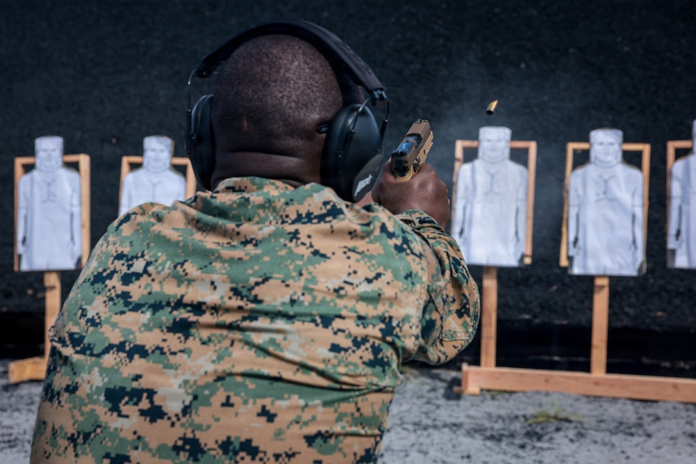 A person in military digital camouflage uniform is firing a semi-automatic pistol at a shooting range. A spent brass casing is visibly ejecting from the firearm. Multiple paper silhouette targets are mounted on wooden stands against a dark backstop.
