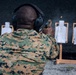 A person in military digital camouflage uniform is firing a semi-automatic pistol at a shooting range. A spent brass casing is visibly ejecting from the firearm. Multiple paper silhouette targets are mounted on wooden stands against a dark backstop.