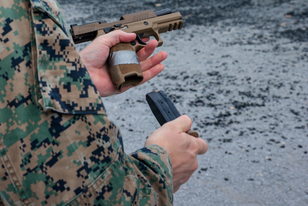 A person wearing USMC MARPAT digital camouflage uniform is performing a magazine change on a flat dark earth (FDE) semi-automatic pistol, consistent with a SIG Sauer P320 M17/M18. The right hand holds the pistol with the magazine well exposed, while the left hand presents a loaded magazine for insertion. The setting appears to be an outdoor shooting range with a gravel or asphalt surface.