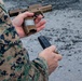 A person wearing USMC MARPAT digital camouflage uniform is performing a magazine change on a flat dark earth (FDE) semi-automatic pistol, consistent with a SIG Sauer P320 M17/M18. The right hand holds the pistol with the magazine well exposed, while the left hand presents a loaded magazine for insertion. The setting appears to be an outdoor shooting range with a gravel or asphalt surface.