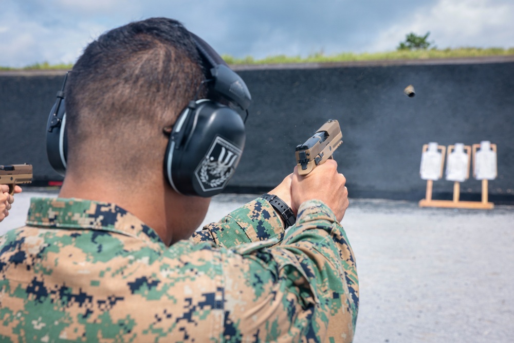 A military or law enforcement personnel is firing a semi-automatic pistol at a shooting range. The shooter is wearing digital camouflage uniform and hearing protection ear muffs. Multiple silhouette targets are visible downrange against a dark backstop wall.