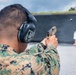 A military or law enforcement personnel is firing a semi-automatic pistol at a shooting range. The shooter is wearing digital camouflage uniform and hearing protection ear muffs. Multiple silhouette targets are visible downrange against a dark backstop wall.