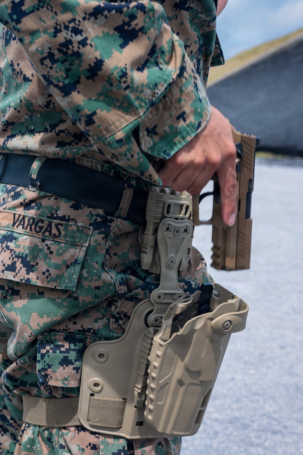 A U.S. Marine in MARPAT woodland digital camouflage uniform is drawing or holstering a pistol at what appears to be a firing range. The name tape 'VARGAS' is clearly visible on the uniform. The Marine is equipped with a tan/coyote brown tactical leg holster mounted on a duty belt.
