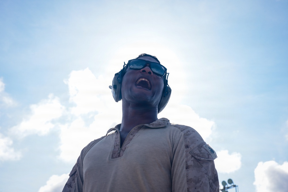 A low-angle photograph of a military personnel wearing desert camouflage uniform, protective sunglasses, and hearing protection earmuffs. The subject is looking upward with mouth open, photographed from below against a bright, partly cloudy sky. The dramatic upward perspective and backlighting create a strong silhouette effect.