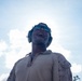 A low-angle photograph of a military personnel wearing desert camouflage uniform, protective sunglasses, and hearing protection earmuffs. The subject is looking upward with mouth open, photographed from below against a bright, partly cloudy sky. The dramatic upward perspective and backlighting create a strong silhouette effect.