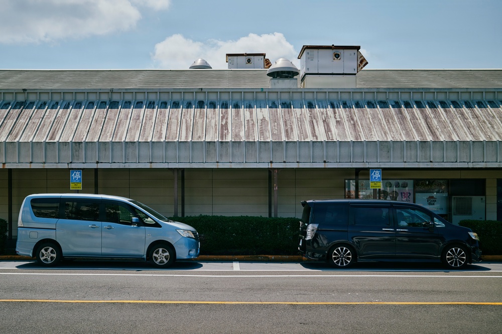 A street-level photograph taken in Japan showing two minivans parked in front of a commercial building with a weathered corrugated metal roof. The left vehicle is light blue and the right is dark navy/black, both appearing to be Japanese domestic market models. Yellow parking signs with the kanji '停' (stop/parking) are visible on the building facade.