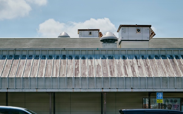 A street-level photograph taken in Japan showing two minivans parked in front of a commercial building with a weathered corrugated metal roof. The left vehicle is light blue and the right is dark navy/black, both appearing to be Japanese domestic market models. Yellow parking signs with the kanji '停' (stop/parking) are visible on the building facade.