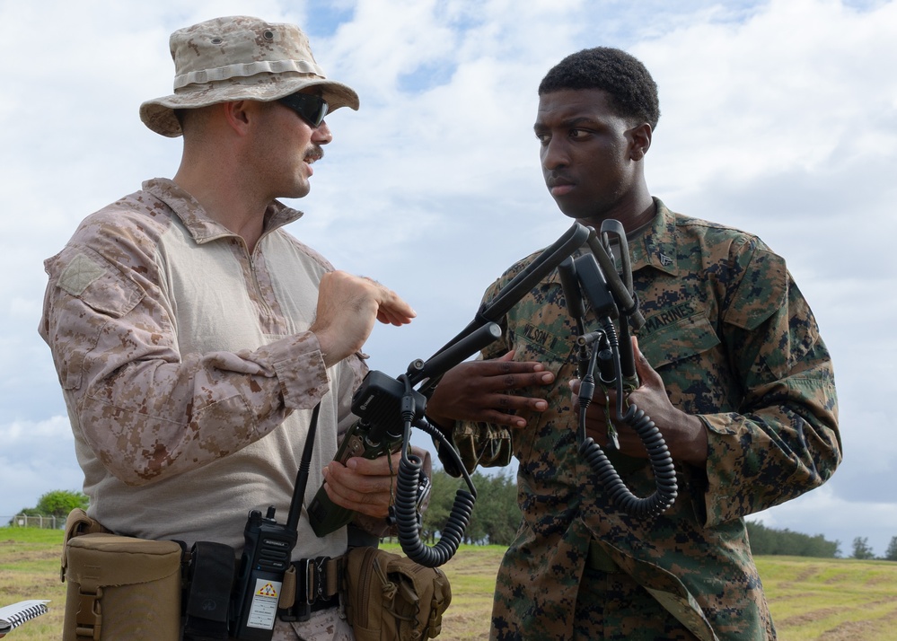Two military personnel in different camouflage uniforms are engaged in what appears to be a training or instructional interaction outdoors. The individual on the left, wearing desert MARPAT camouflage and a boonie hat, appears to be instructing the Marine on the right, who is wearing woodland MARPAT and holding communication equipment with multiple antenna connectors. The setting appears to be an open field with a partly cloudy sky in the background.