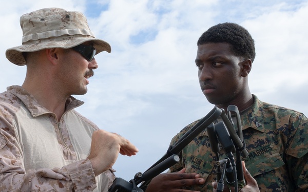 Two military personnel in different camouflage uniforms are engaged in what appears to be a training or instructional interaction outdoors. The individual on the left, wearing desert MARPAT camouflage and a boonie hat, appears to be instructing the Marine on the right, who is wearing woodland MARPAT and holding communication equipment with multiple antenna connectors. The setting appears to be an open field with a partly cloudy sky in the background.