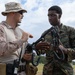 Two military personnel in different camouflage uniforms are engaged in what appears to be a training or instructional interaction outdoors. The individual on the left, wearing desert MARPAT camouflage and a boonie hat, appears to be instructing the Marine on the right, who is wearing woodland MARPAT and holding communication equipment with multiple antenna connectors. The setting appears to be an open field with a partly cloudy sky in the background.