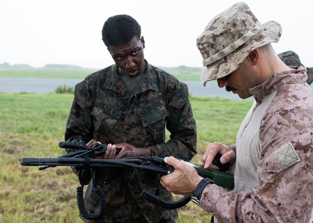 Two military personnel in camouflage uniforms are examining or handling communications or tactical equipment in an outdoor field setting. The individual on the right wears desert MARPAT camouflage with a boonie hat, while the individual on the left wears woodland MARPAT camouflage. They appear to be conducting equipment inspection or instruction in overcast, possibly rainy conditions.