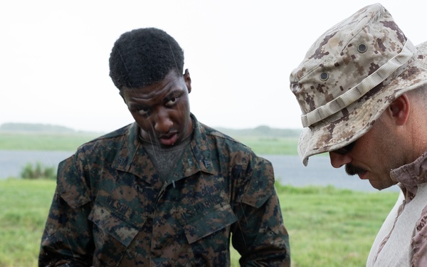Two military personnel in camouflage uniforms are examining or handling communications or tactical equipment in an outdoor field setting. The individual on the right wears desert MARPAT camouflage with a boonie hat, while the individual on the left wears woodland MARPAT camouflage. They appear to be conducting equipment inspection or instruction in overcast, possibly rainy conditions.