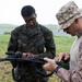 Two military personnel in camouflage uniforms are examining or handling communications or tactical equipment in an outdoor field setting. The individual on the right wears desert MARPAT camouflage with a boonie hat, while the individual on the left wears woodland MARPAT camouflage. They appear to be conducting equipment inspection or instruction in overcast, possibly rainy conditions.