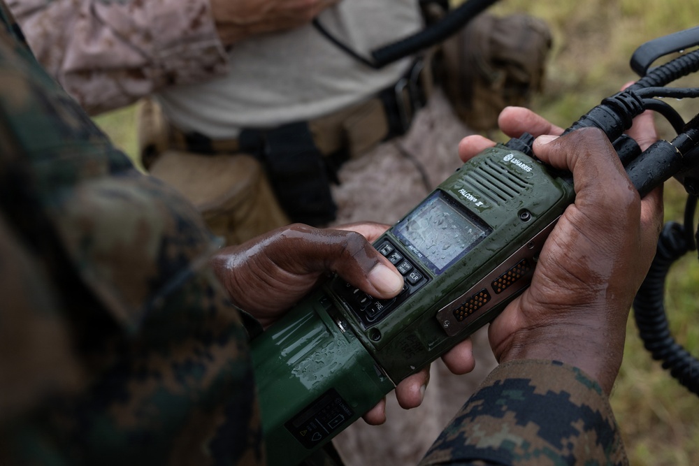A person in military camouflage uniform is holding a green L3Harris Falcon III military tactical radio, which appears wet from rain or moisture. The device is being operated or configured in what appears to be a field environment. A second individual in mixed civilian/military attire is visible in the background.