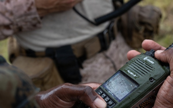 A person in military camouflage uniform is holding a green L3Harris Falcon III military tactical radio, which appears wet from rain or moisture. The device is being operated or configured in what appears to be a field environment. A second individual in mixed civilian/military attire is visible in the background.