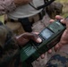 A person in military camouflage uniform is holding a green L3Harris Falcon III military tactical radio, which appears wet from rain or moisture. The device is being operated or configured in what appears to be a field environment. A second individual in mixed civilian/military attire is visible in the background.