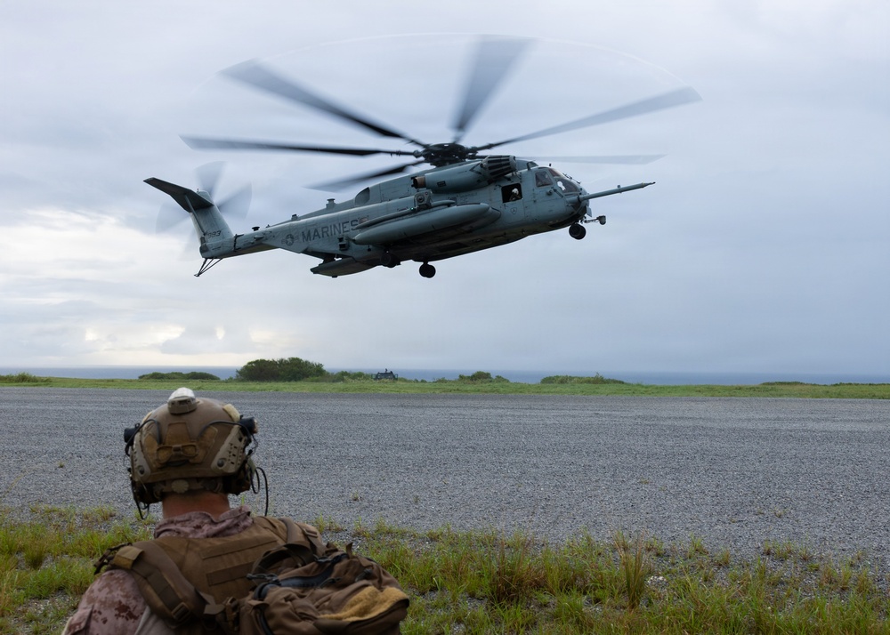 A U.S. Marines CH-53E Super Stallion heavy-lift helicopter is hovering or landing on a gravel landing zone near a coastal area under overcast skies. A Marine in full combat gear, including a tactical helmet with communications equipment and a backpack, is observing the aircraft from a kneeling or prone position in the foreground. The scene appears to depict a military training exercise or tactical insertion/extraction operation.