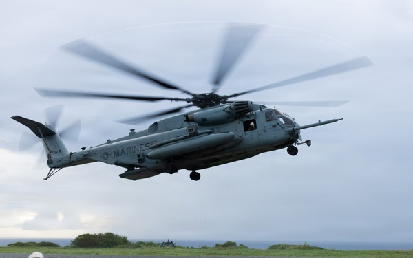 A U.S. Marines CH-53E Super Stallion heavy-lift helicopter is hovering or landing on a gravel landing zone near a coastal area under overcast skies. A Marine in full combat gear, including a tactical helmet with communications equipment and a backpack, is observing the aircraft from a kneeling or prone position in the foreground. The scene appears to depict a military training exercise or tactical insertion/extraction operation.
