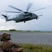 A U.S. Marines CH-53E Super Stallion heavy-lift helicopter is hovering or landing on a gravel landing zone near a coastal area under overcast skies. A Marine in full combat gear, including a tactical helmet with communications equipment and a backpack, is observing the aircraft from a kneeling or prone position in the foreground. The scene appears to depict a military training exercise or tactical insertion/extraction operation.