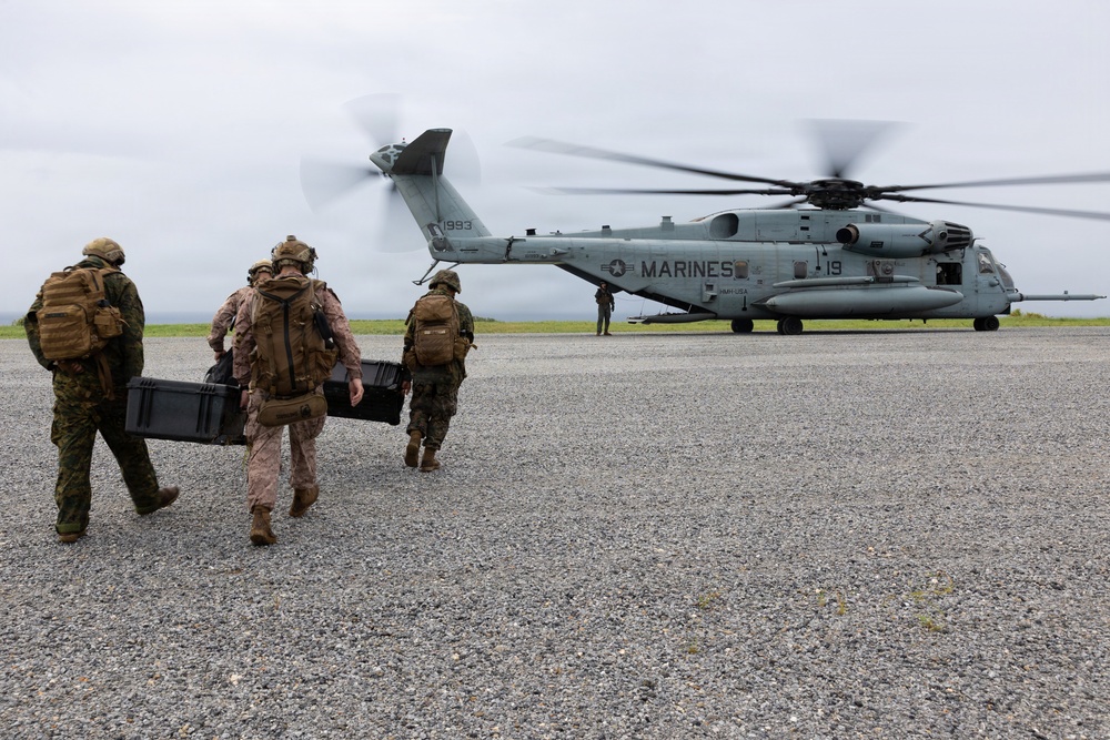 Military personnel, including U.S. Marines and what appears to be allied forces in different camouflage patterns, are approaching a large CH-53E Super Stallion helicopter marked 'MARINES' with tail number 1993. The personnel are carrying equipment cases and wearing full tactical gear including helmets and backpacks. The scene takes place on a gravel landing zone under overcast skies.