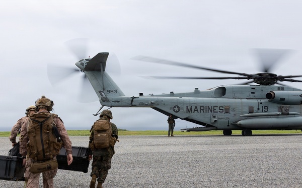 Military personnel, including U.S. Marines and what appears to be allied forces in different camouflage patterns, are approaching a large CH-53E Super Stallion helicopter marked 'MARINES' with tail number 1993. The personnel are carrying equipment cases and wearing full tactical gear including helmets and backpacks. The scene takes place on a gravel landing zone under overcast skies.