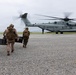 Military personnel, including U.S. Marines and what appears to be allied forces in different camouflage patterns, are approaching a large CH-53E Super Stallion helicopter marked 'MARINES' with tail number 1993. The personnel are carrying equipment cases and wearing full tactical gear including helmets and backpacks. The scene takes place on a gravel landing zone under overcast skies.