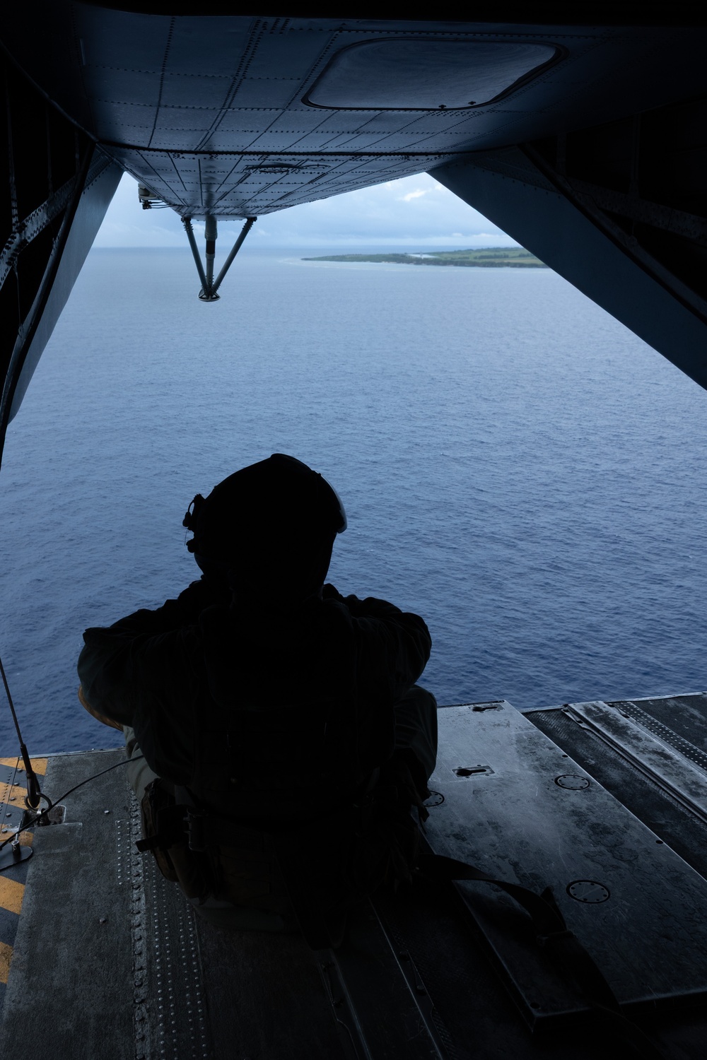 A silhouetted military aircrew member sits on the open rear ramp of a military transport aircraft, looking out over a blue ocean with a small island or atoll visible in the distance. The view is taken from inside the aircraft looking outward through the open cargo ramp. The scene suggests an aerial reconnaissance, airdrop preparation, or observation mission over a tropical maritime environment.
