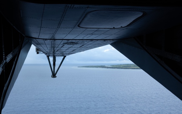 A silhouetted military aircrew member sits on the open rear ramp of a military transport aircraft, looking out over a blue ocean with a small island or atoll visible in the distance. The view is taken from inside the aircraft looking outward through the open cargo ramp. The scene suggests an aerial reconnaissance, airdrop preparation, or observation mission over a tropical maritime environment.