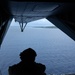 A silhouetted military aircrew member sits on the open rear ramp of a military transport aircraft, looking out over a blue ocean with a small island or atoll visible in the distance. The view is taken from inside the aircraft looking outward through the open cargo ramp. The scene suggests an aerial reconnaissance, airdrop preparation, or observation mission over a tropical maritime environment.
